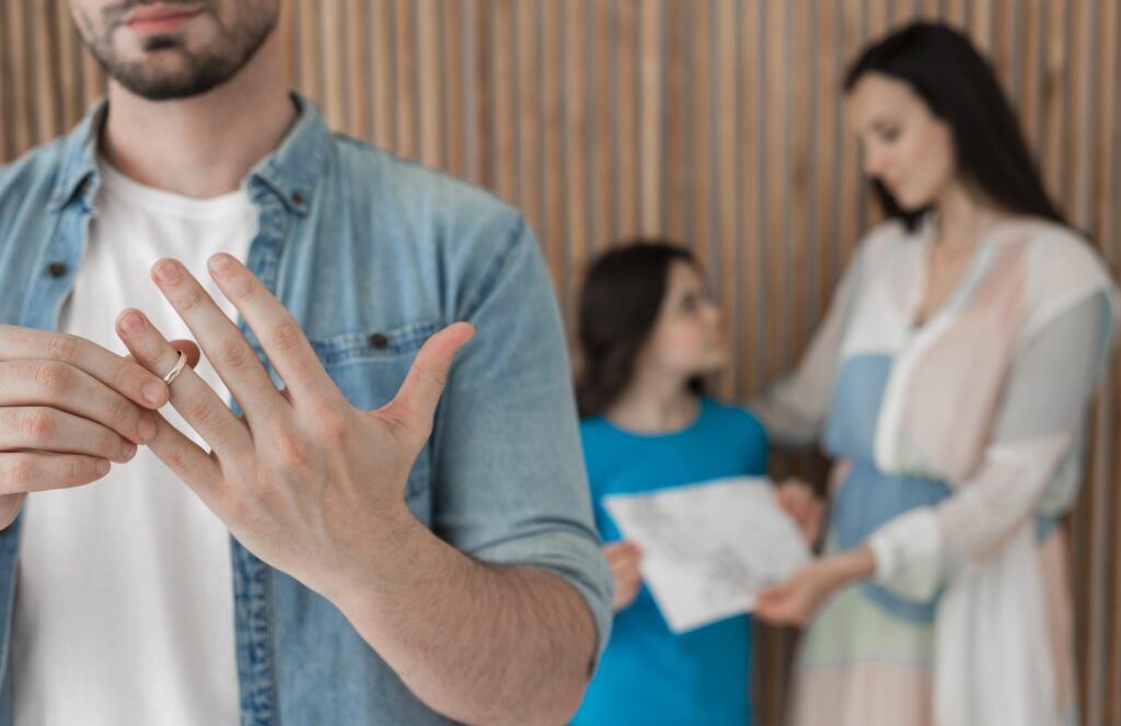 A man removing his wedding ring while his family watches in the background, symbolizing emotional strain and raising the question is codependence a result of divorced parents.