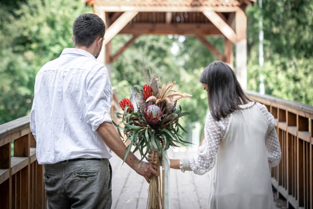 Back view of a couple holding an exotic bouquet during their vow renewal ceremony on a wooden bridge.