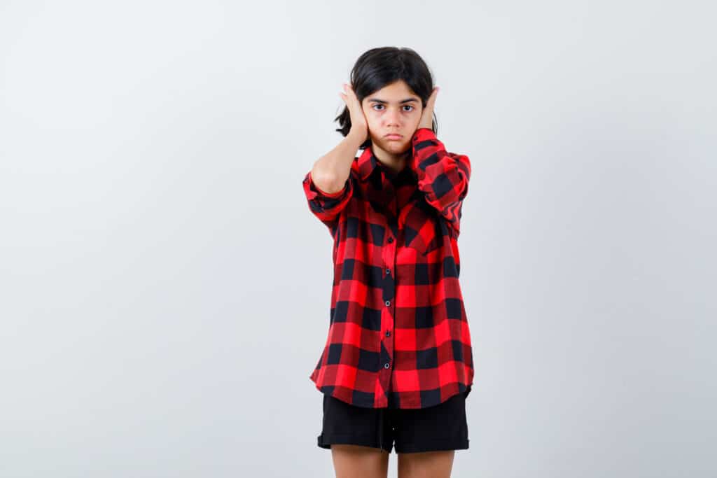 Disobedient Child Outline: A young girl in a red plaid shirt covering her ears with a defiant expression, illustrating the challenges of managing disobedient behavior in children.