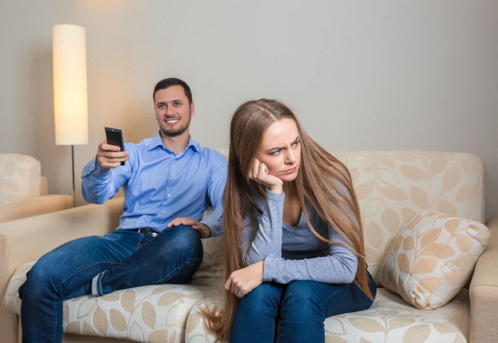 A couple sitting on a sofa; the man, smiling, holds a remote control, while the woman looks frustrated. This image represents a common situation where learning how to communicate when upset can improve relationship dynamics.