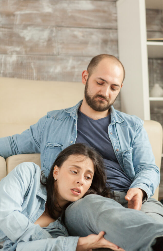 A tired woman leaning on her partner's lap, both parents appearing reflective and concerned, illustrating the emotional toll of constant struggles in parenting