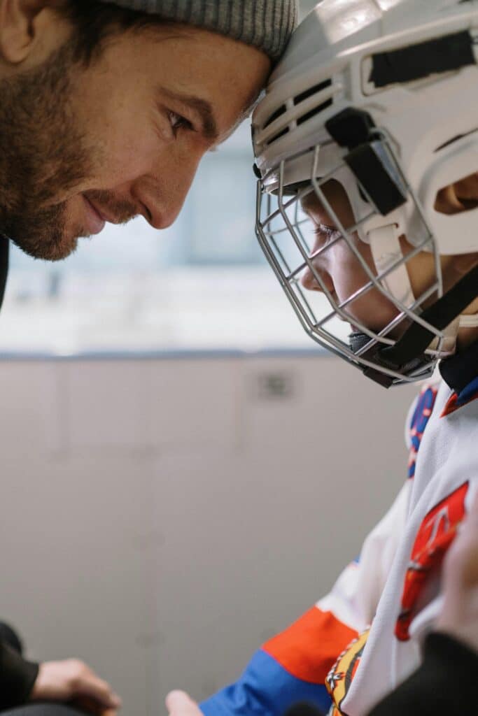 A father leans in closely, looking into his child's eyes with encouragement. The child, dressed in a hockey uniform and helmet, listens intently. A moment of motivation and support before the game. What motivates your child to push through challenges?